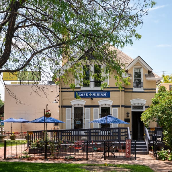 Cafe Miriam's yellow building with blue trim, outdoor patio seating with umbrellas, and green trees.