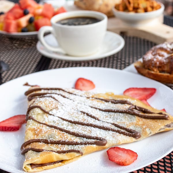 A chocolate-drizzled crepe with powdered sugar and strawberries, coffee, and fruit salad on a table.
