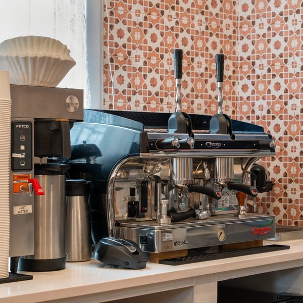 A Fetco coffee machine and a chrome Astoria espresso machine on a white counter with patterned tiles.