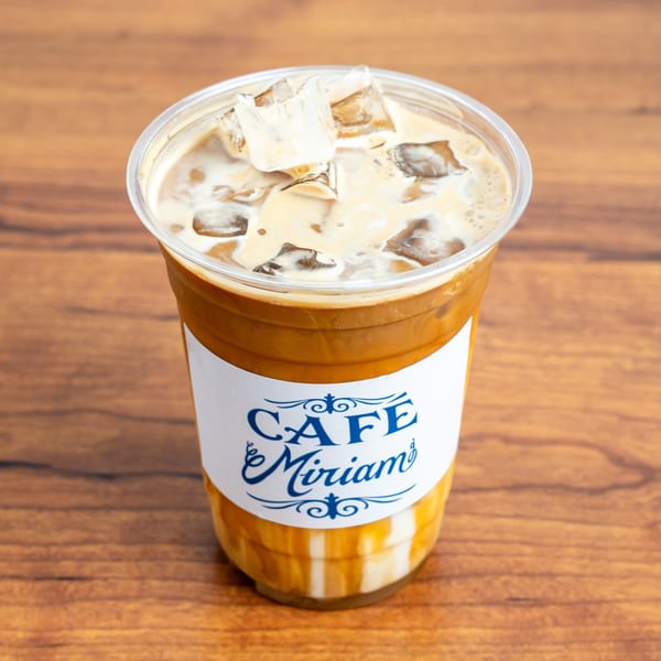 Iced coffee with cream and ice in a clear Cafe Miriam cup on a wooden table.