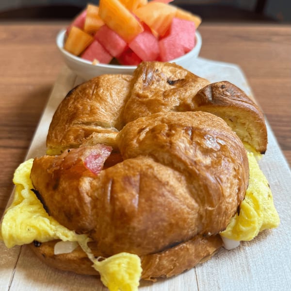 A croissant breakfast sandwich with scrambled eggs and bacon, served with a side of fruit.