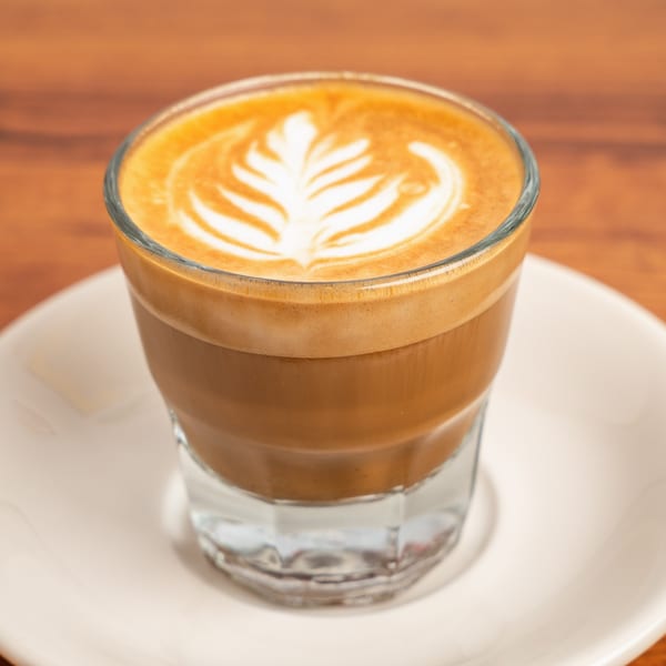A Cafe Miriam latte in a clear glass with intricate leaf-shaped latte art on a white saucer.