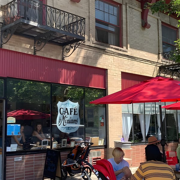 Cafe Miriam's exterior with red awnings, outdoor seating under red umbrellas, and people dining.