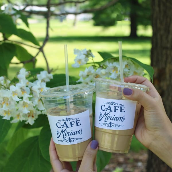 Two hands holding Cafe Miriam iced coffees with white blossoms and green foliage in the background.