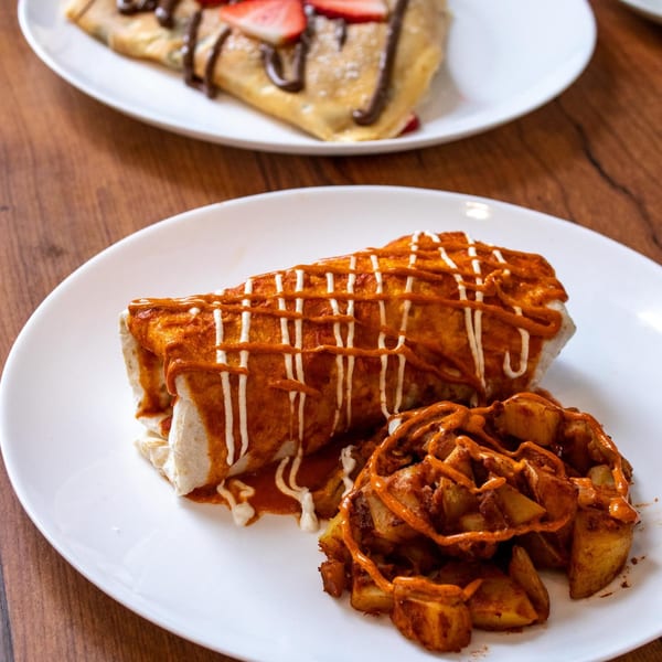 A breakfast burrito with red and white sauce, served with seasoned potato wedges on a white plate.
