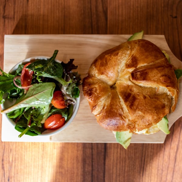 Croissant sandwich with avocado and egg, served with a side salad on a wooden board.