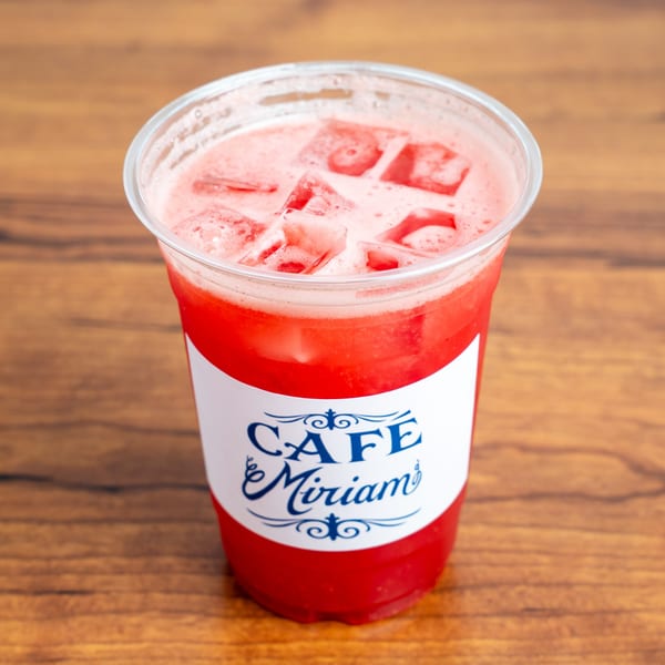 Cafe Miriam's iced strawberry drink with foam and ice cubes, on a wooden table.