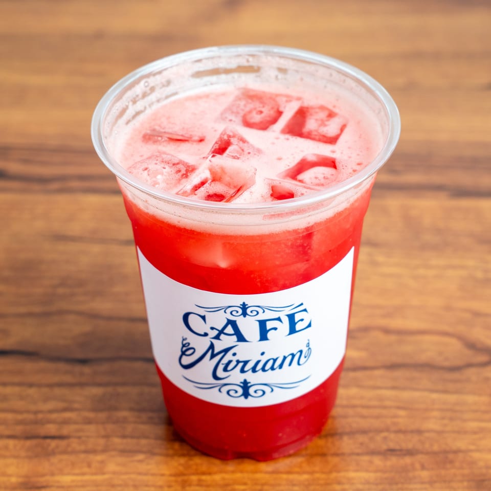 Cafe Miriam's iced strawberry drink with foam and ice cubes, on a wooden table.