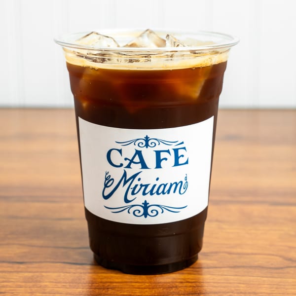 Iced coffee in a clear plastic cup with a "Cafe Miriam" label on a wooden table.