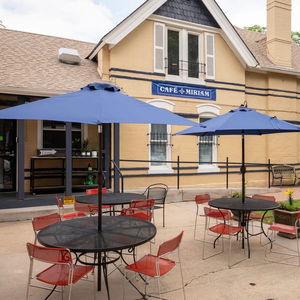 Cafe Miriam's outdoor patio features black tables, red chairs, and blue umbrellas in front of a yellow building.