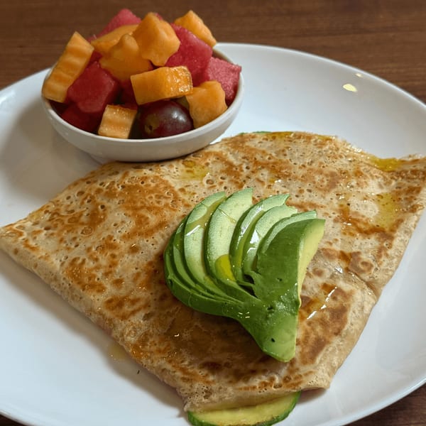 A breakfast crepe topped with sliced avocado and honey, served with a side of fresh fruit.